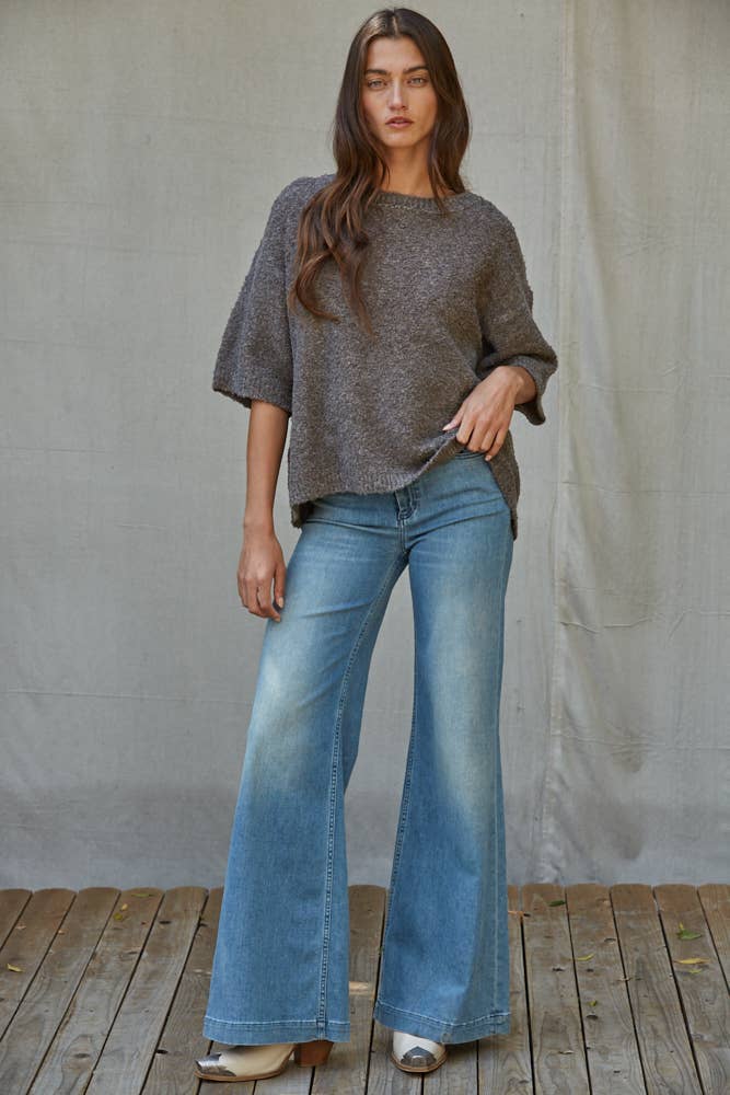 A woman modeling a loose, short-sleeve heathered gray knit top and light blue wide-leg jeans, standing on a wooden plank floor against a neutral draped fabric backdrop.