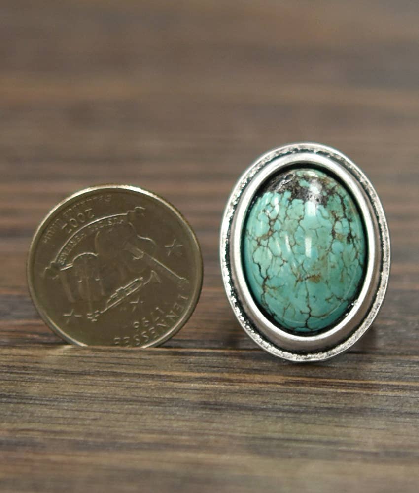 A silver ring with a large oval turquoise stone, shown next to a US quarter coin for scale on a dark wood surface.