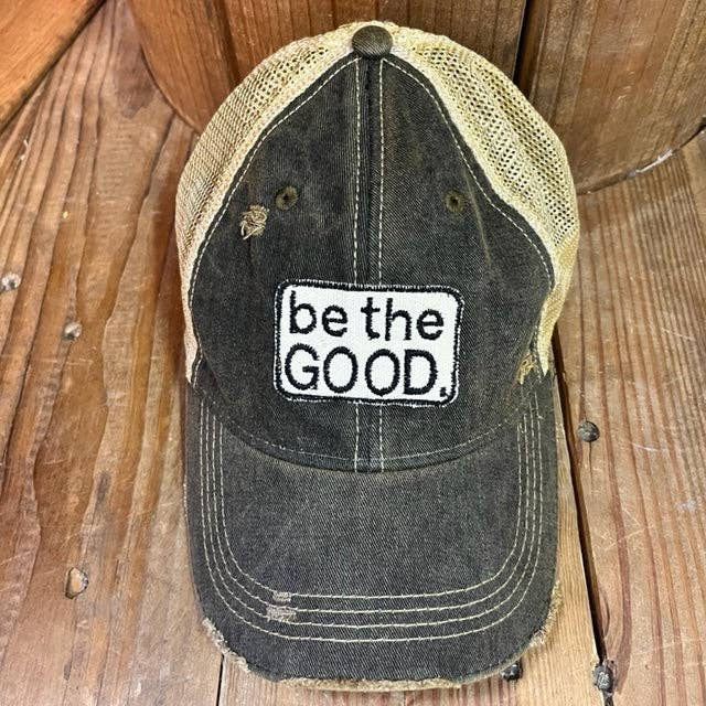 Black and beige cap with 'be the GOOD.' text on a wooden surface
