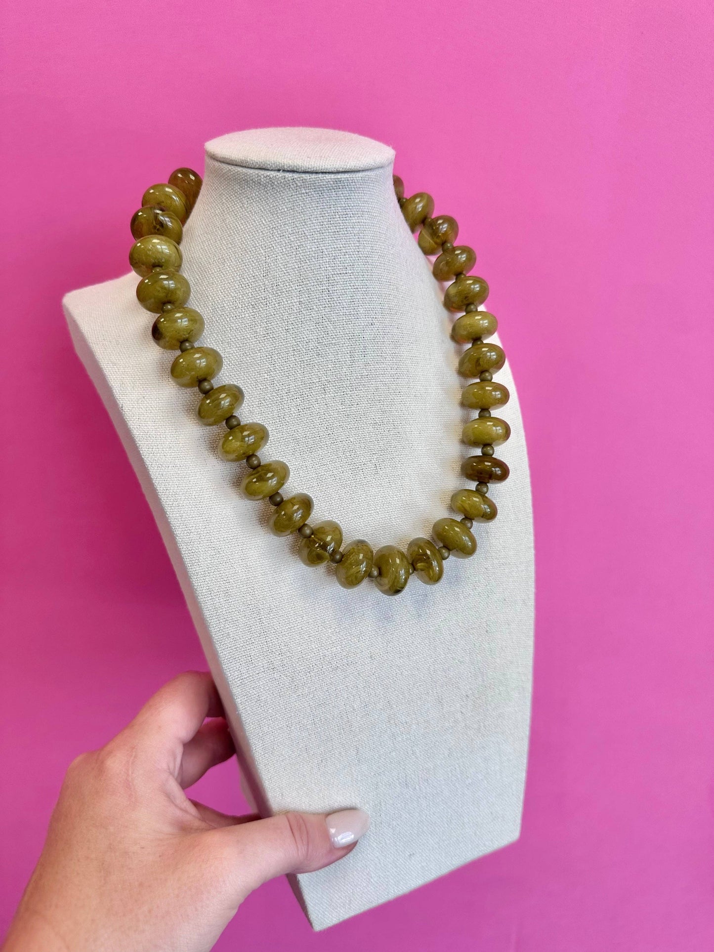 A chunky olive green beaded necklace displayed on a light gray fabric mannequin bust, held at the bottom by a hand against a solid pink background.