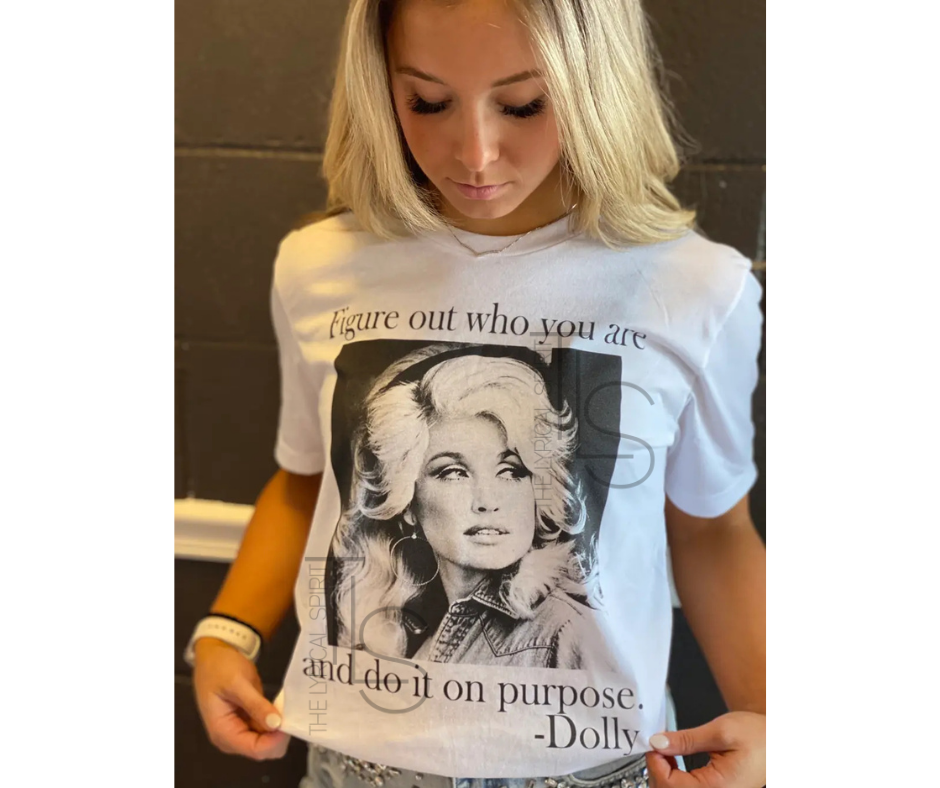 A woman wearing a white graphic t-shirt featuring a black-and-white portrait of Dolly Parton with the quote 'Figure out who you are and do it on purpose. -Dolly'.