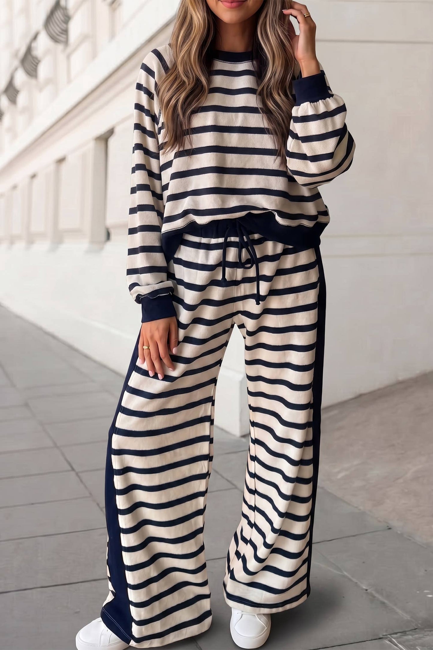 A woman wearing a matching two-piece loungewear set with navy blue and cream horizontal stripes, standing on a sidewalk next to a white building.