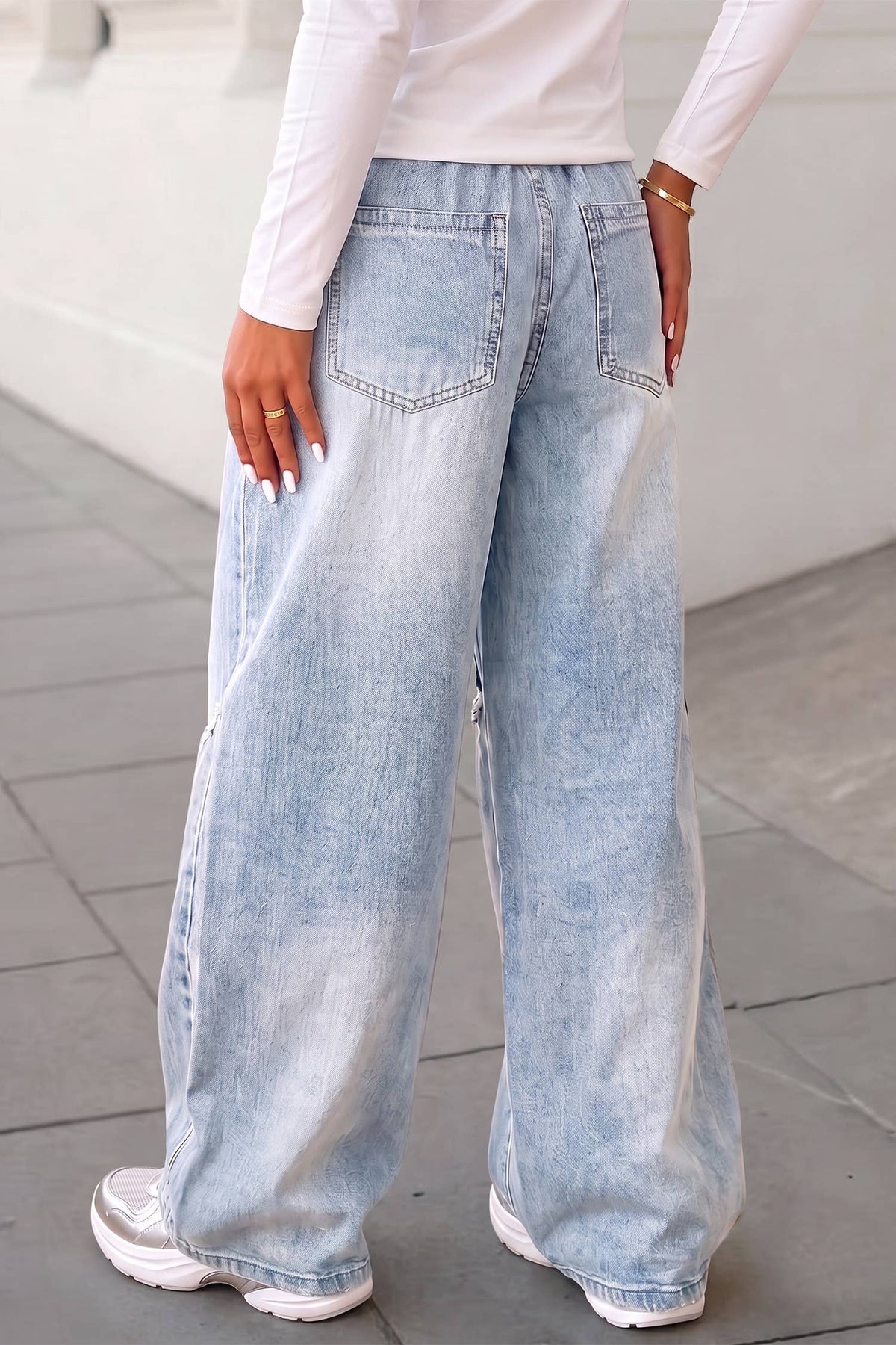 A person shown from the back wearing very wide-leg light-wash blue jeans with a faded wash, a white long-sleeve top, and white sneakers, standing on a gray tiled floor.