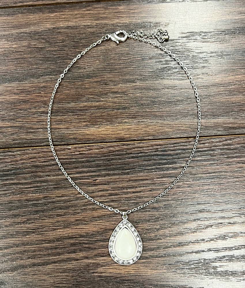 A silver-toned chain necklace with a teardrop-shaped pendant featuring a white inlay and a border of small clear stones, displayed on a dark wood surface.