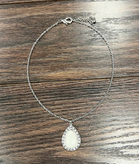 A silver-toned chain necklace with a teardrop-shaped pendant featuring a white inlay and a border of small clear stones, displayed on a dark wood surface.