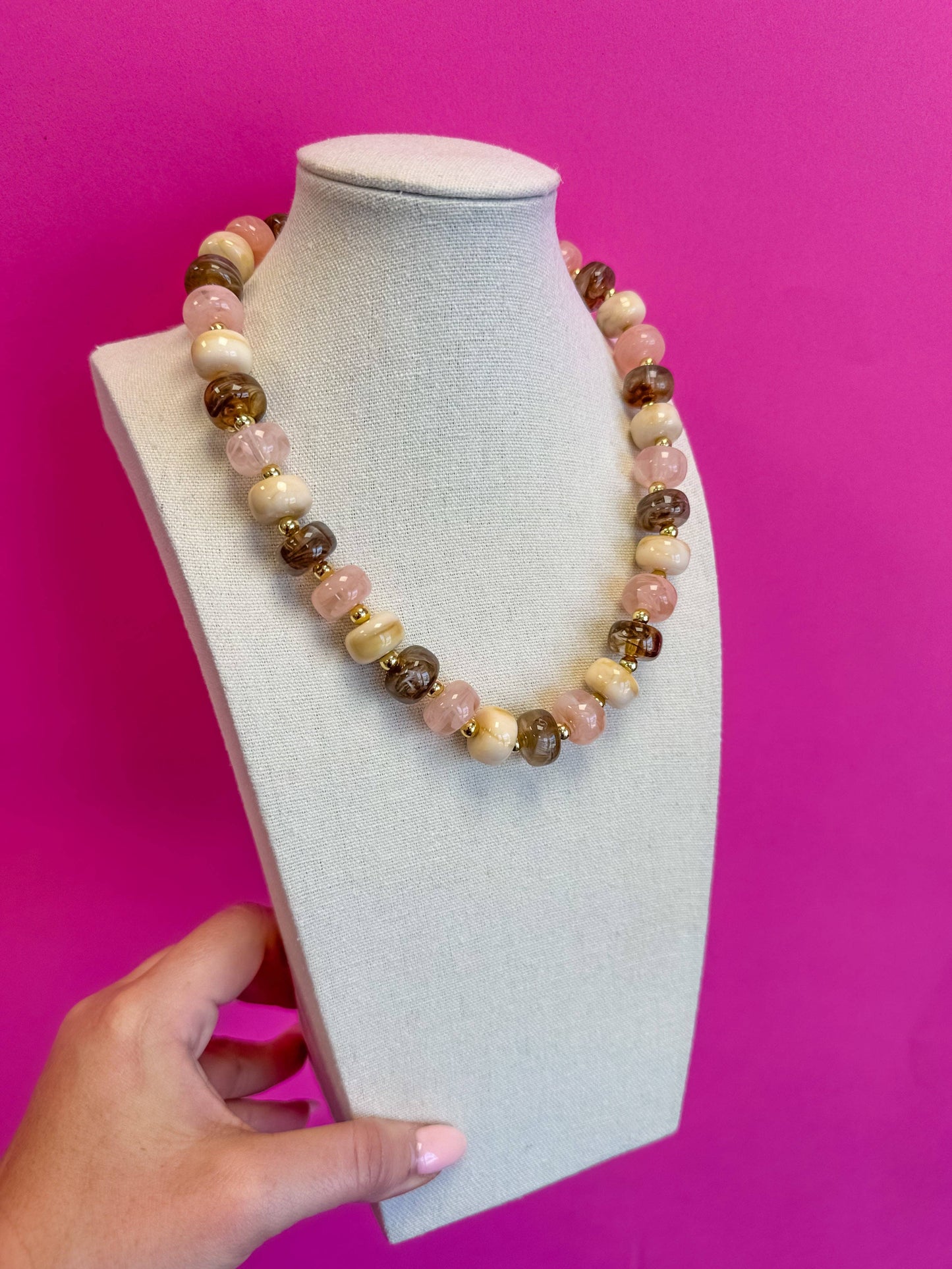 A beaded necklace with pink, cream, and brown gemstone-like beads displayed on a light gray fabric mannequin bust, with a hand holding the base against a bright pink background.