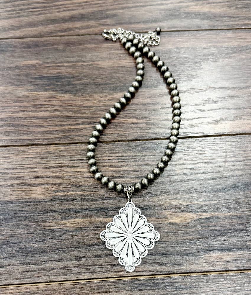 A beaded necklace with dark metallic beads and a large silver-toned sunburst pendant, displayed on a dark wood background.