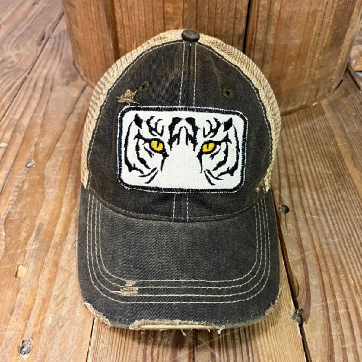 A distressed trucker-style baseball cap with a dark washed front panel, tan mesh back, and a white embroidered tiger face patch with yellow eyes on the front. The cap is photographed on a rustic wooden surface with a wooden barrel in the background.