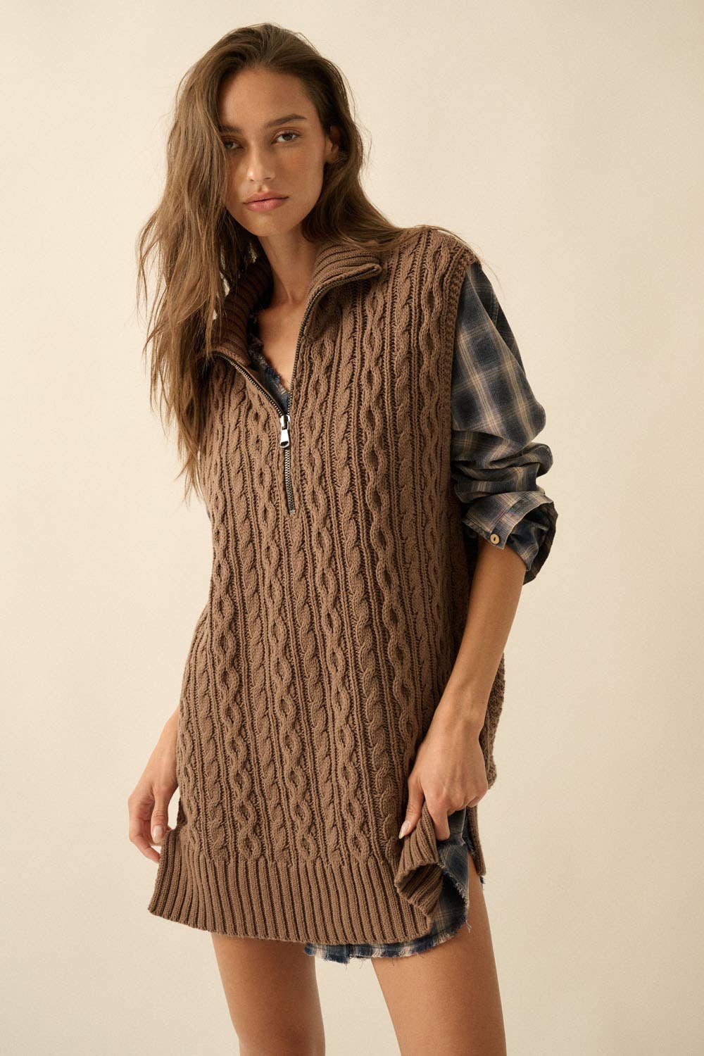 Woman wearing a brown cable knit vest over a plaid shirt on a beige background