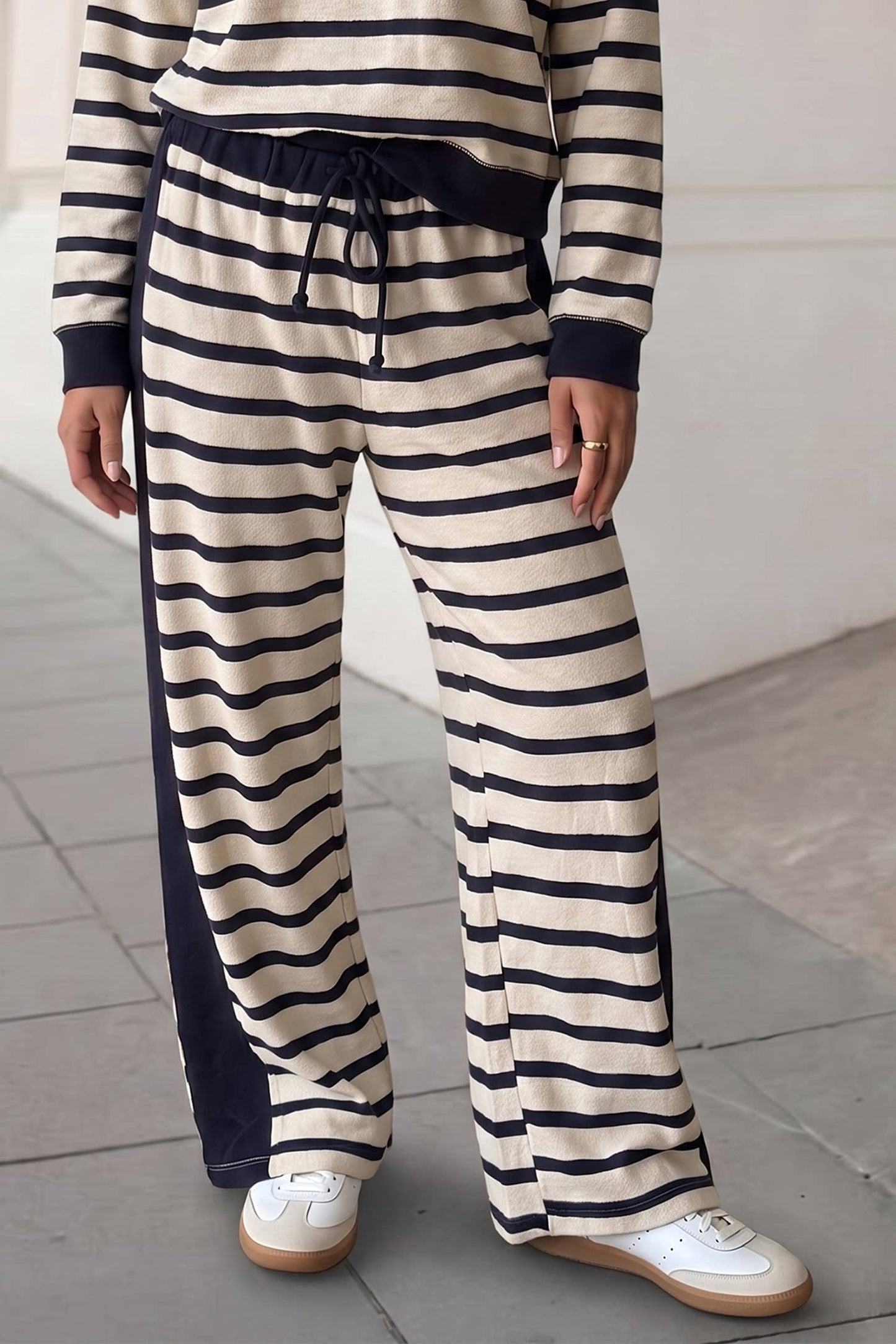 A person wearing loose striped knit pants with navy side panels and a navy drawstring waist, paired with white sneakers, photographed indoors against a light wall and tiled floor.