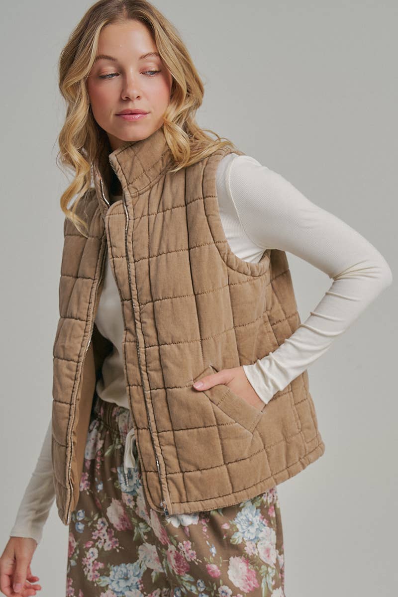 A woman wearing a tan quilted puffer vest over a white long-sleeve top and a floral skirt, photographed against a plain light gray studio background.