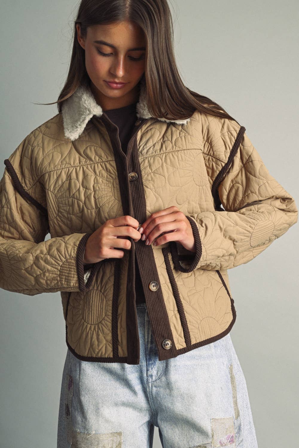 A woman wearing a beige quilted jacket with dark brown trim and a white sherpa collar, buttoning up the front. She is also wearing light wash jeans against a plain light gray studio background.