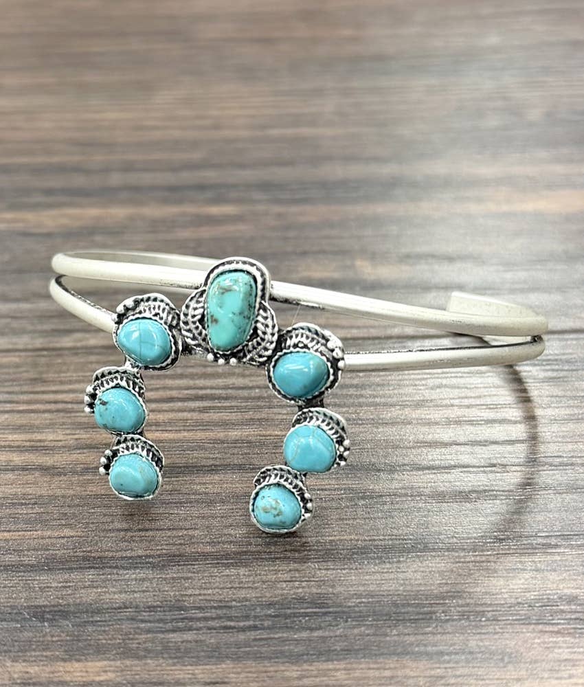 A silver cuff bracelet decorated with multiple turquoise stones in an ornate Southwestern style, resting on a dark wood surface.