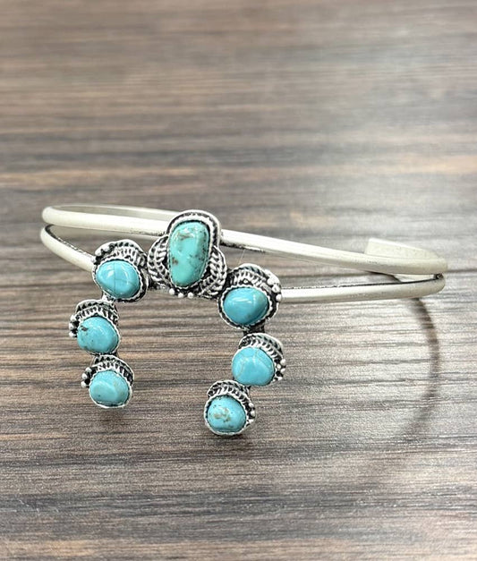 A silver cuff bracelet decorated with multiple turquoise stones in an ornate Southwestern style, resting on a dark wood surface.