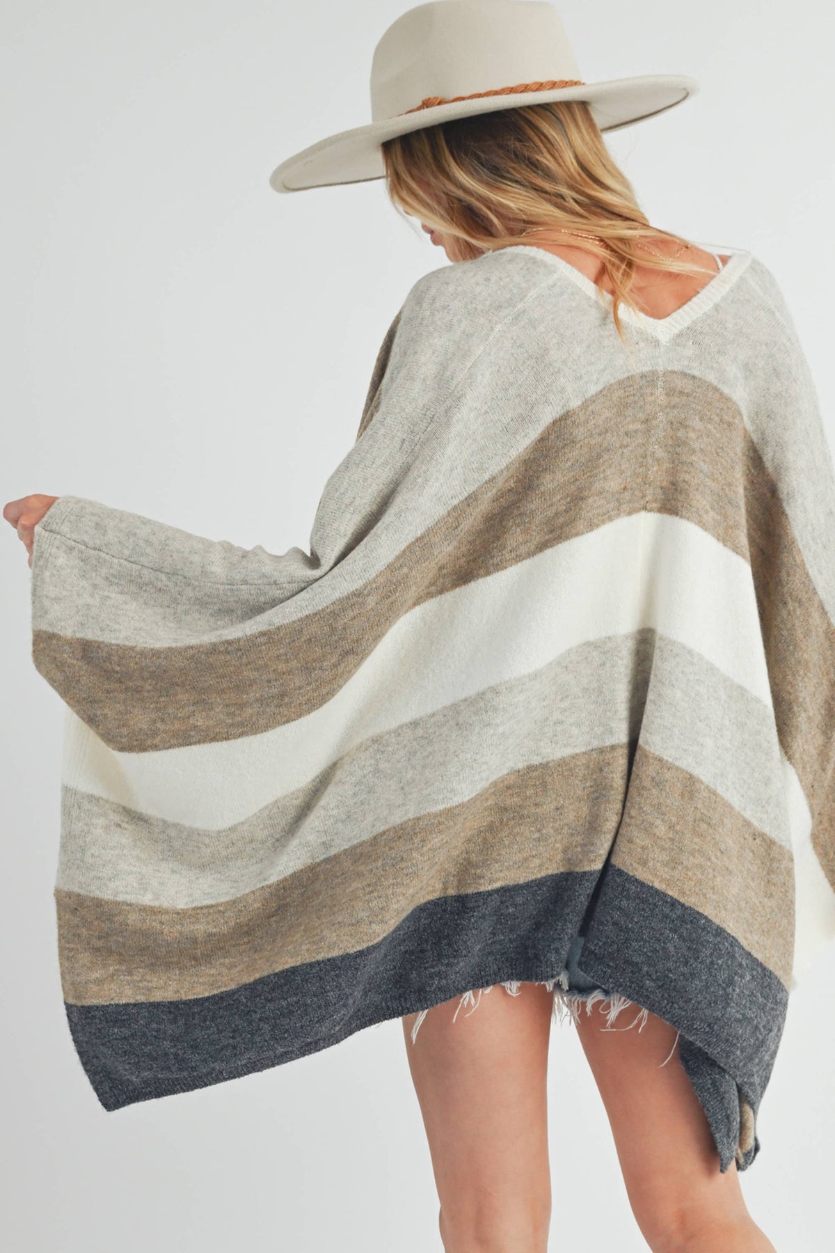 A woman shown from the back wearing a wide-brim beige hat and a loose striped knit poncho with horizontal bands of cream, taupe, beige, and charcoal gray on a light gray studio background.