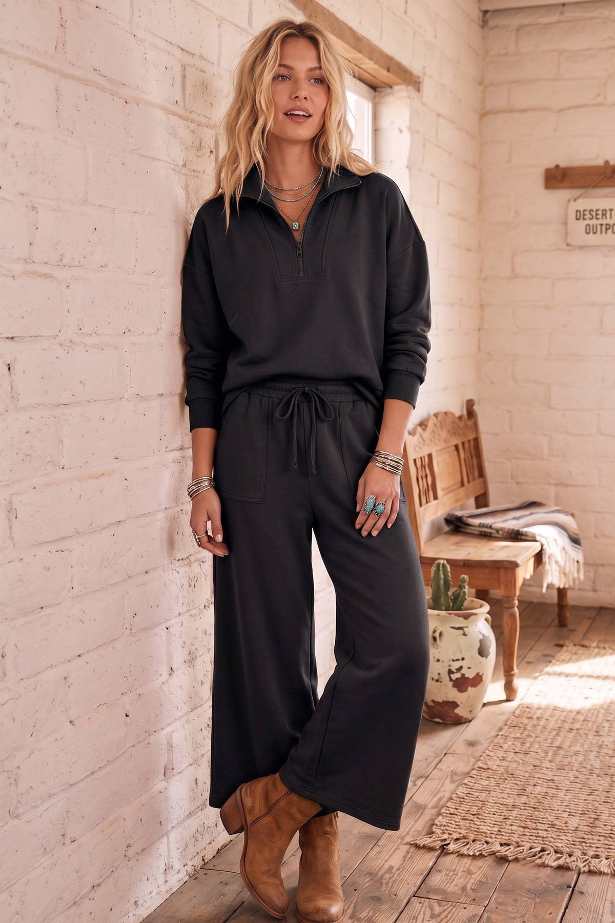A woman stands in a rustic indoor setting wearing a black long-sleeve quarter-zip pullover and matching wide-leg drawstring pants, paired with tan ankle boots.