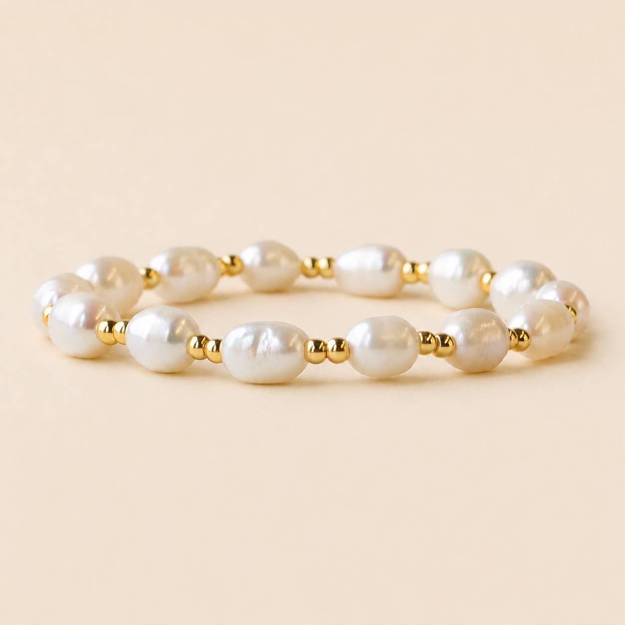 A beaded stretch bracelet featuring irregular white freshwater pearls separated by small gold spacer beads, displayed on a solid light beige background.