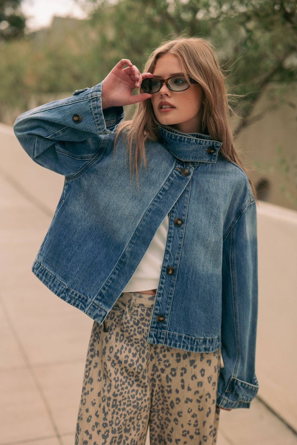 A woman wearing a cropped blue denim jacket over a white top and leopard-print pants, standing outdoors on a paved walkway with trees in the background.