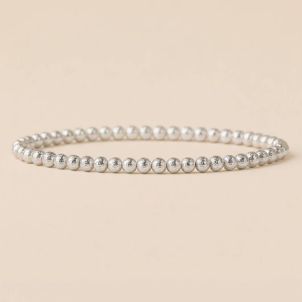 A silver beaded stretch bracelet with small, polished metallic beads, displayed on a solid beige background.