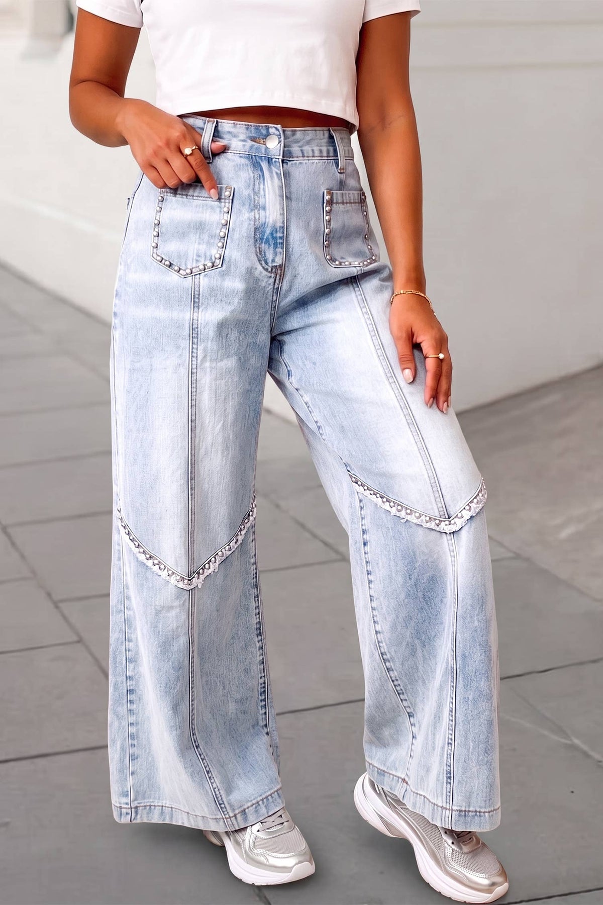 A person wearing light-wash wide-leg denim jeans with decorative silver chain trim along the front pockets and upper thighs, paired with a white cropped top and white sneakers.