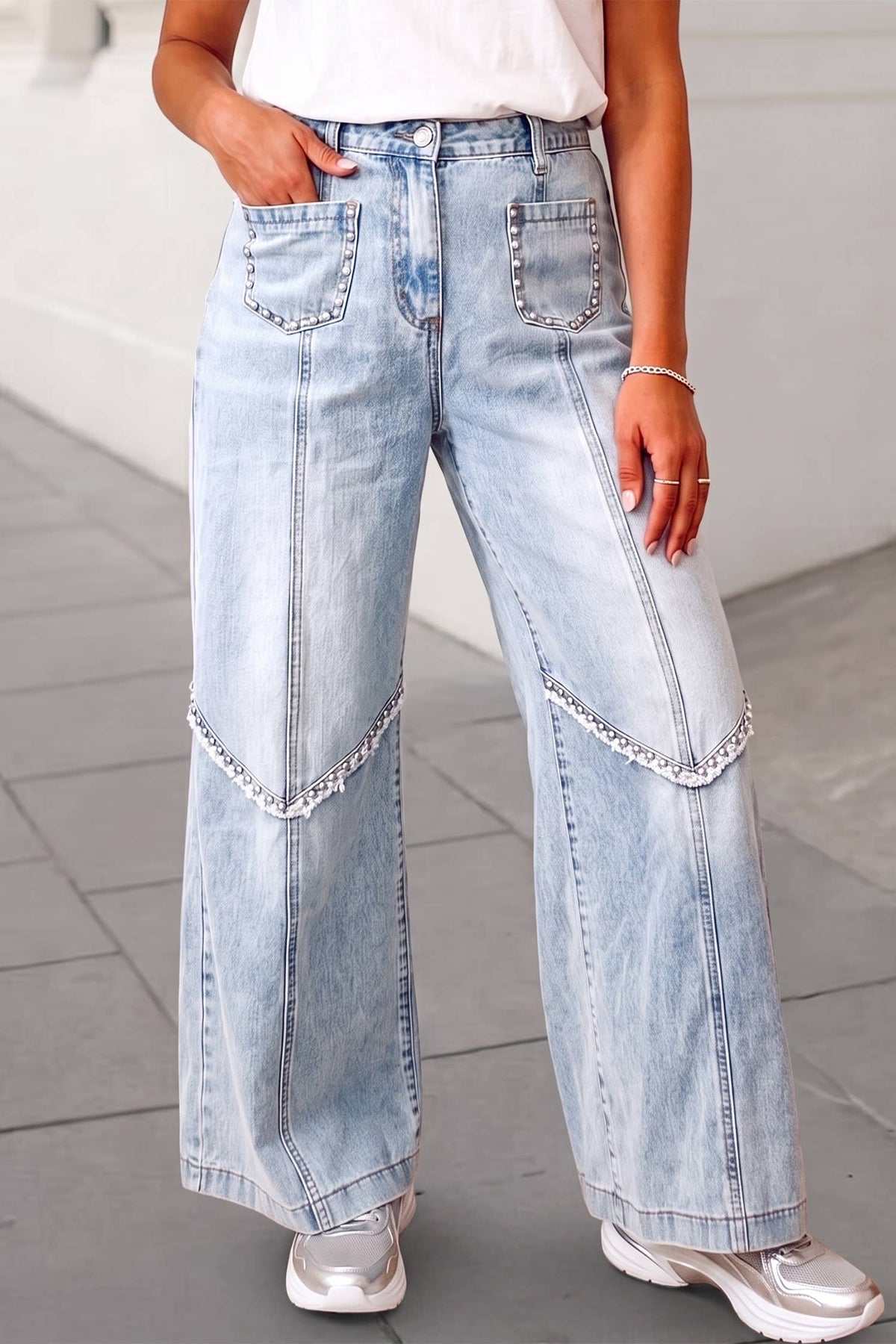 A person wearing light-wash wide-leg denim jeans with decorative silver chain embellishments on the front of the legs, paired with a white top and silver sneakers.