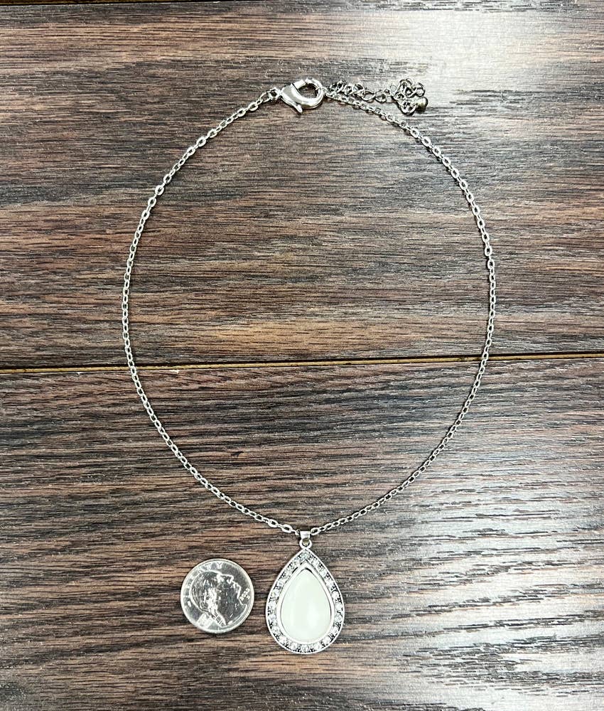 A silver chain necklace with a teardrop-shaped white stone pendant and a coin pendant, displayed on a dark wood background.