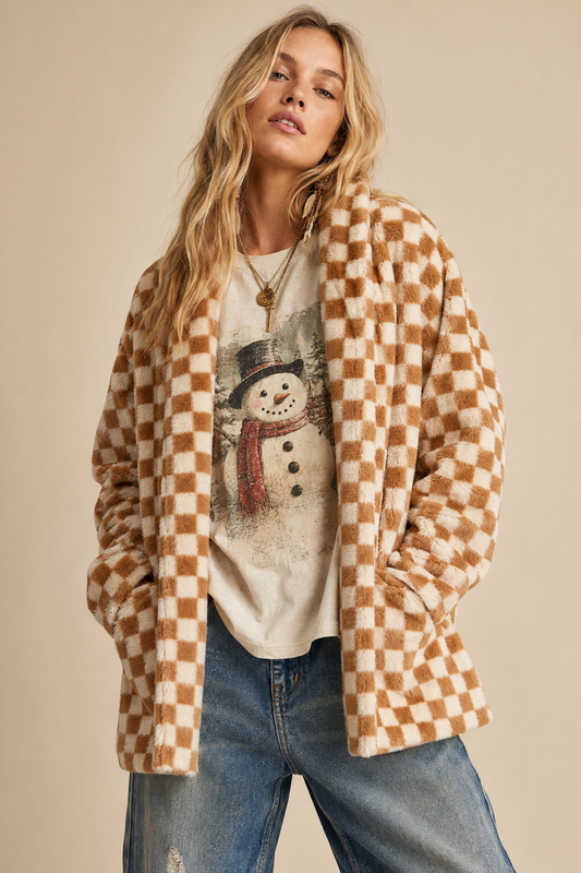A woman wearing a brown and beige checkered fuzzy cardigan over a cream graphic t-shirt and blue jeans, posed against a solid beige background.