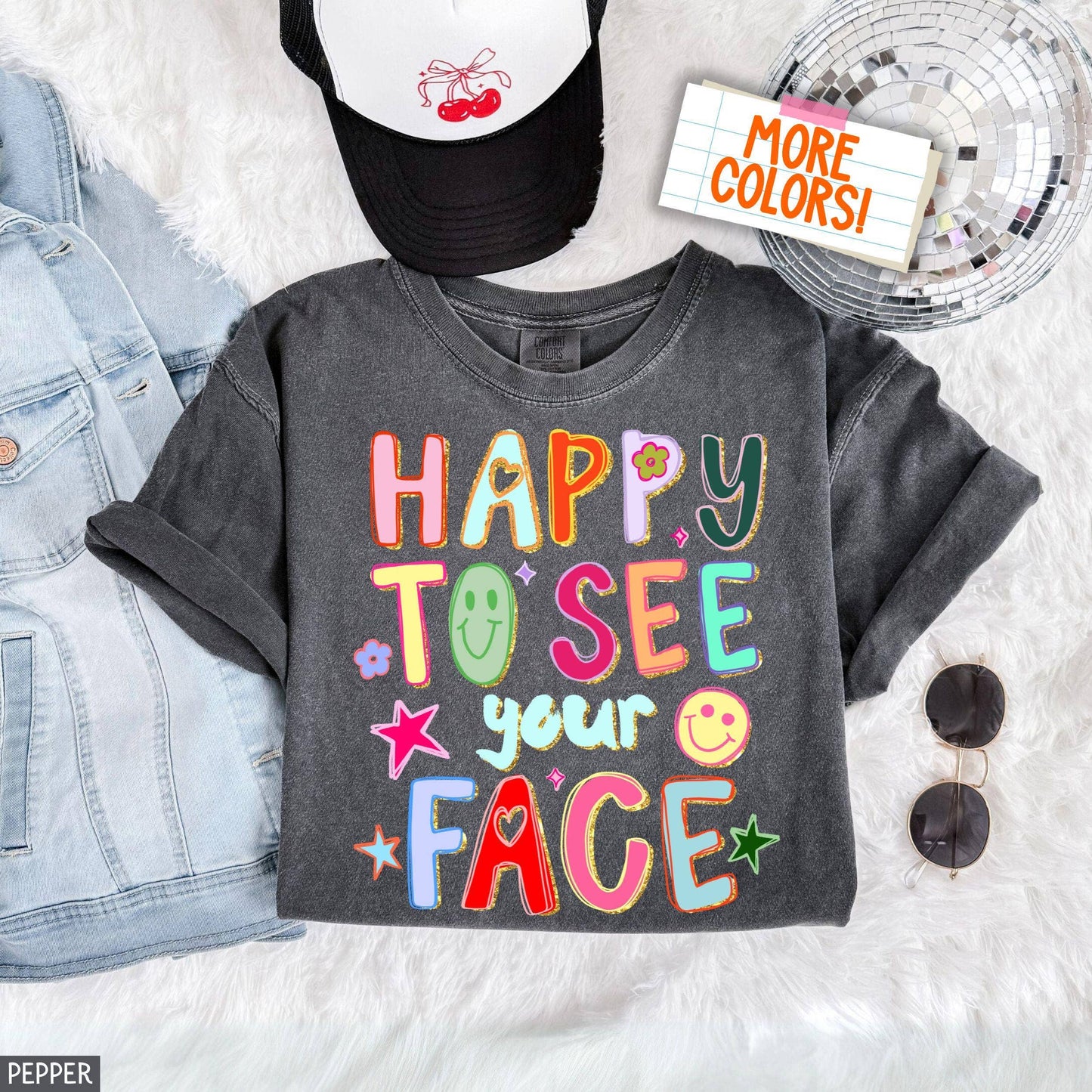 Gray t-shirt with colorful text 'Happy to see your face' on a light gray background with denim jacket, sunglasses, and cap.