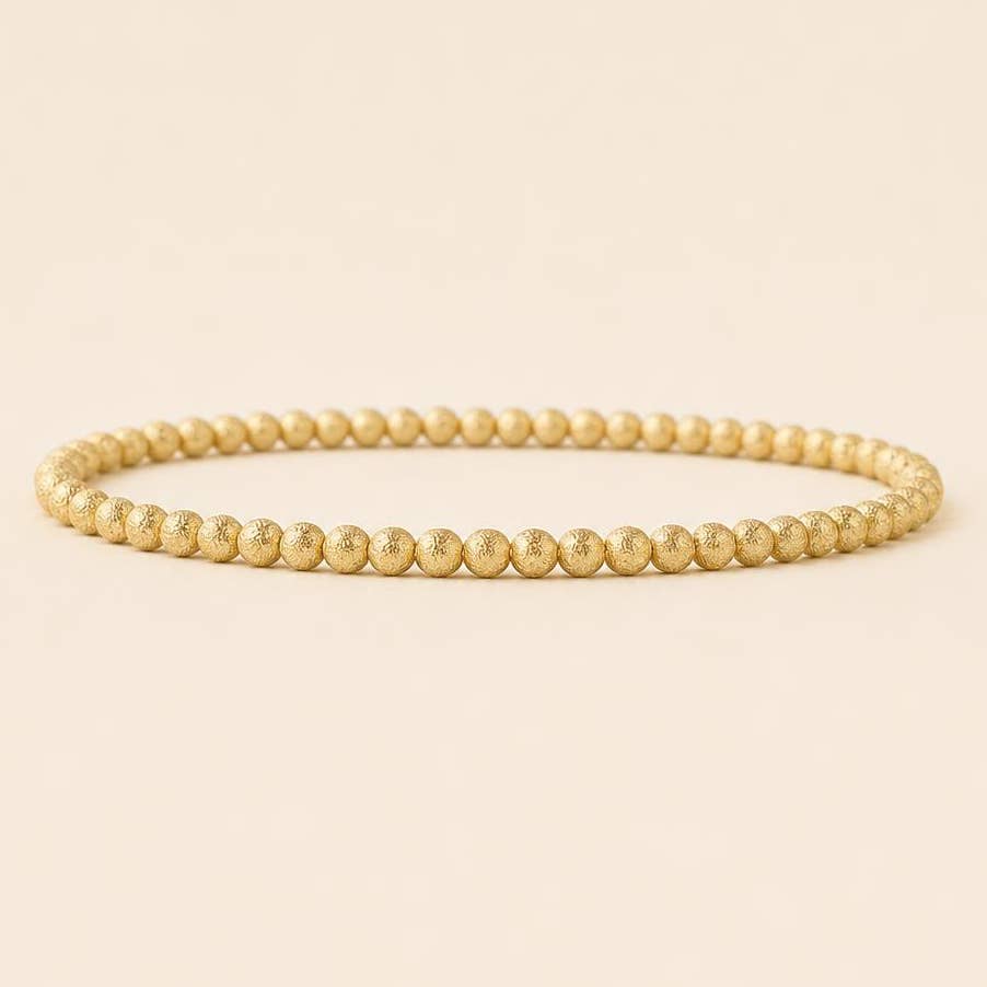 A single gold beaded bracelet with a textured, matte finish, arranged in a horizontal circle on a solid, warm beige background.