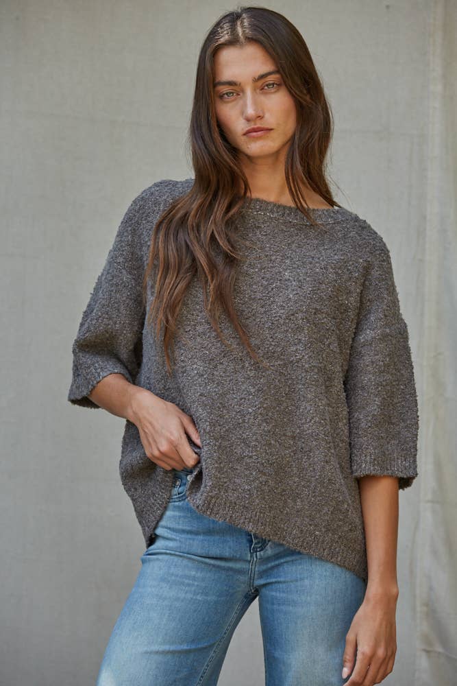 A woman wearing a loose, heathered gray knit sweater with three-quarter sleeves and blue jeans, standing against a neutral concrete-textured wall.