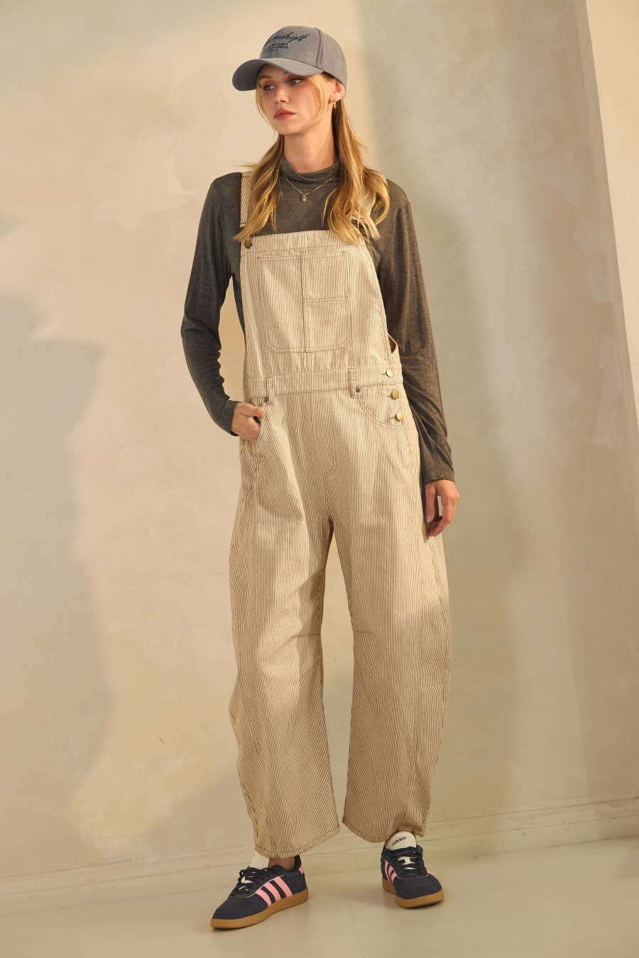 Woman wearing beige overalls and a gray cap against a neutral background