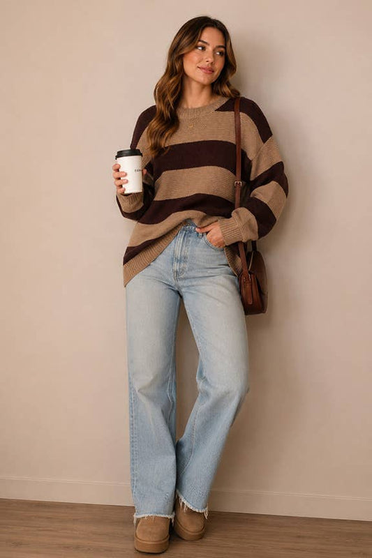 A woman stands against a beige wall wearing a brown and beige horizontally striped knit sweater, light-wash blue jeans, and tan boots, while holding a white coffee cup and carrying a brown shoulder bag.