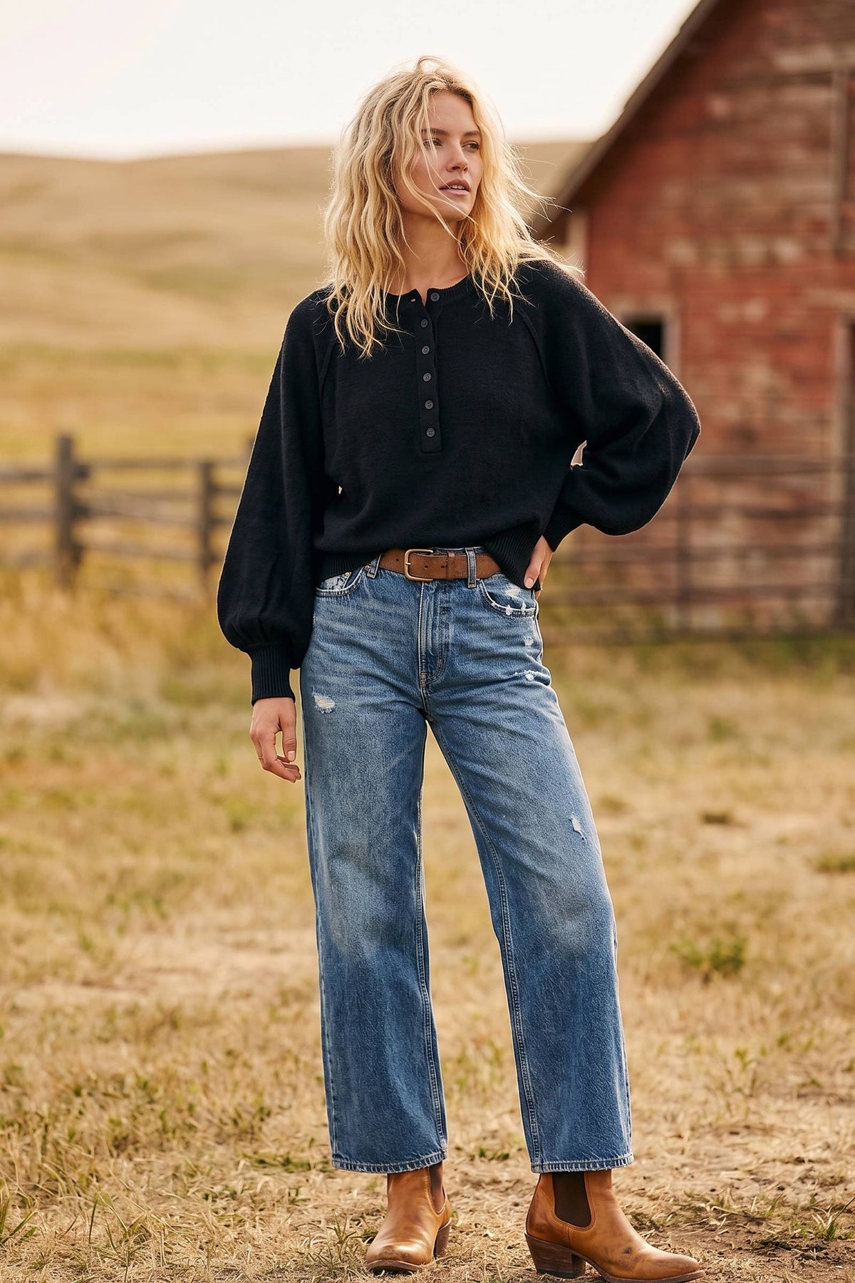 Woman wearing a black sweater and blue jeans standing in a field with a barn in the background