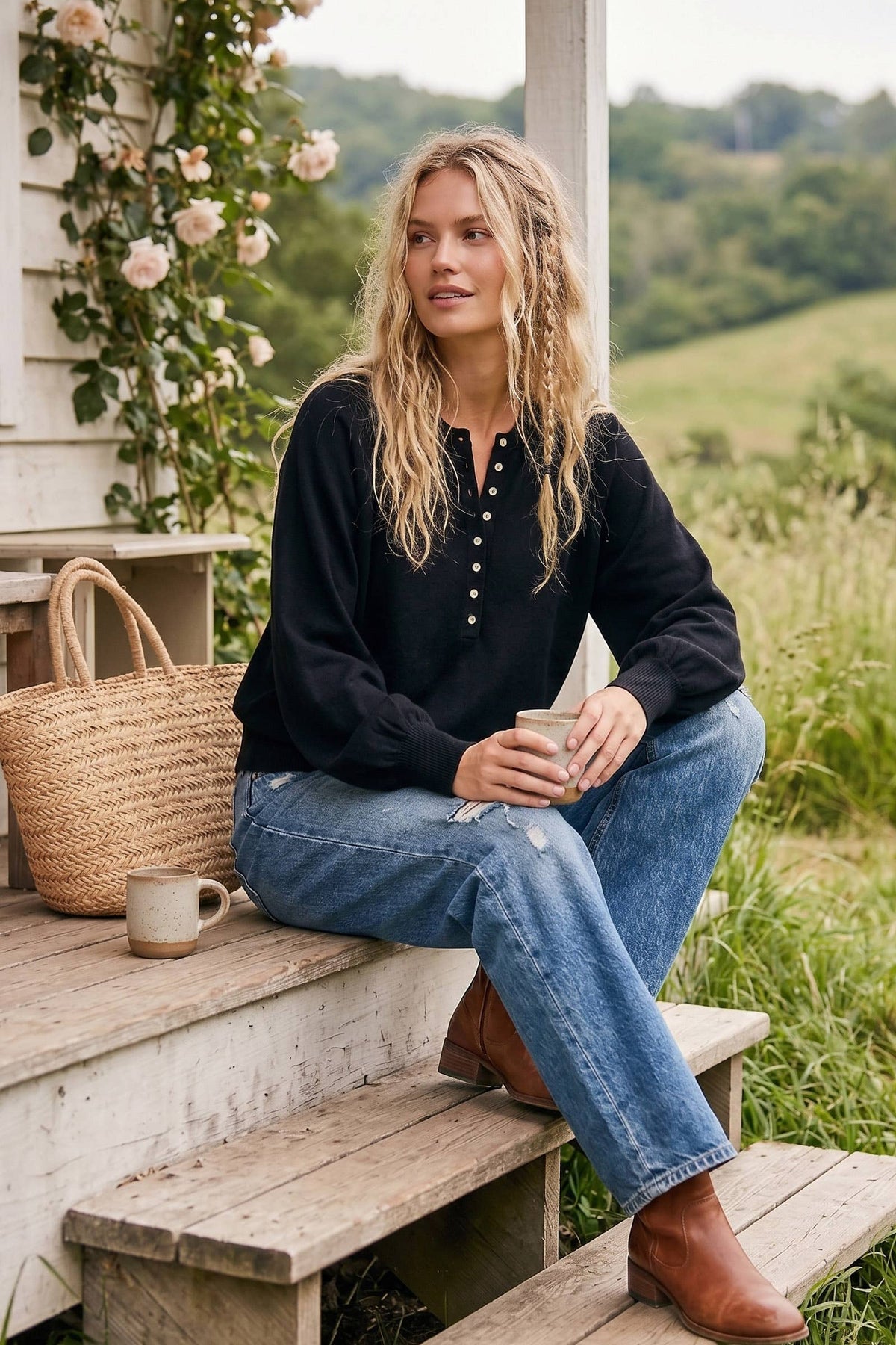 A woman sits on rustic wooden porch steps wearing a black long-sleeve henley top with blue jeans and brown ankle boots, holding a mug in an outdoor garden setting.