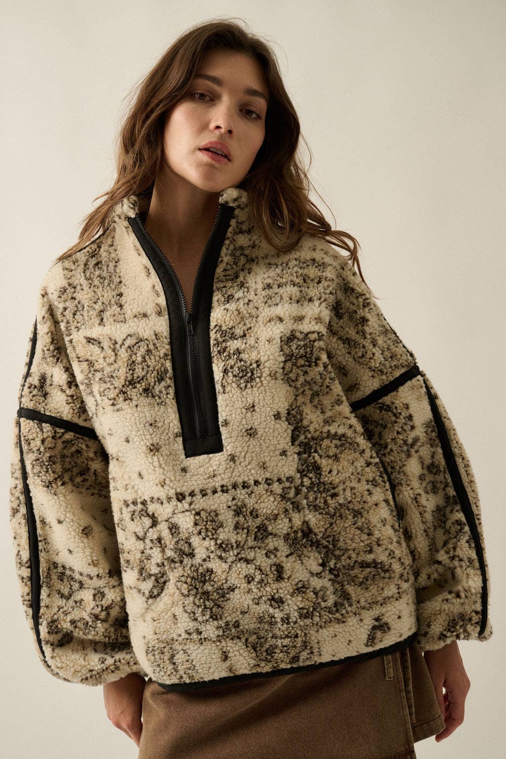 A woman wearing a cream and brown patterned sherpa pullover with black trim and a black zipper, photographed against a plain light beige studio background.