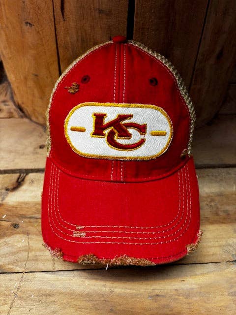 A distressed red Kansas City Chiefs baseball cap with a white and gold embroidered 'KC' logo patch on the front, resting on a rustic wooden surface.
