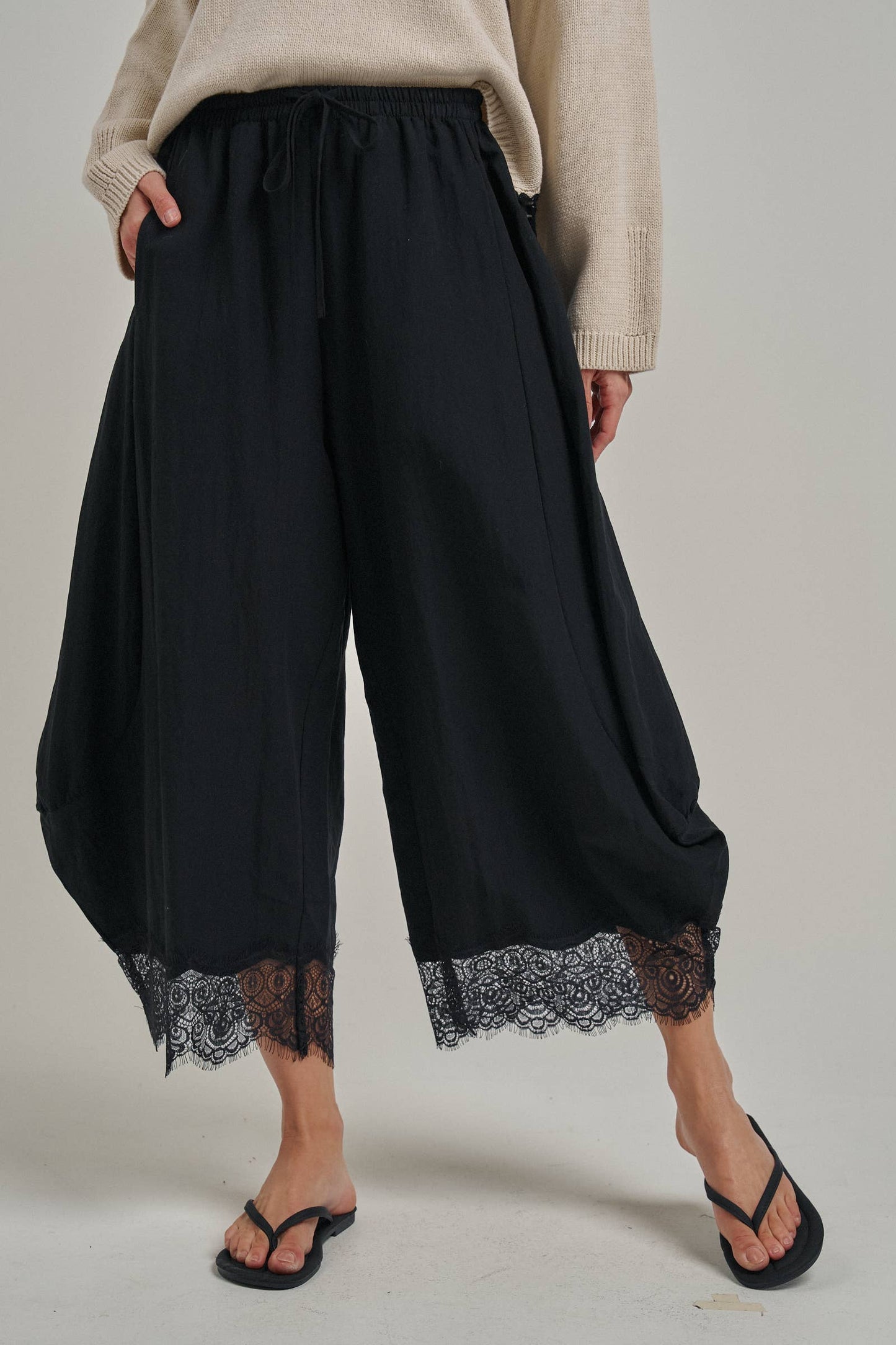 Black wide-leg cropped pants with a lace hem, worn by a model with a beige sweater and black sandals against a light gray studio background.