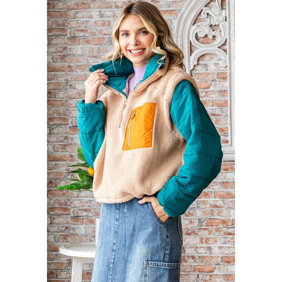 Woman wearing a beige puffer vest with orange pocket and teal sleeves against a brick wall.