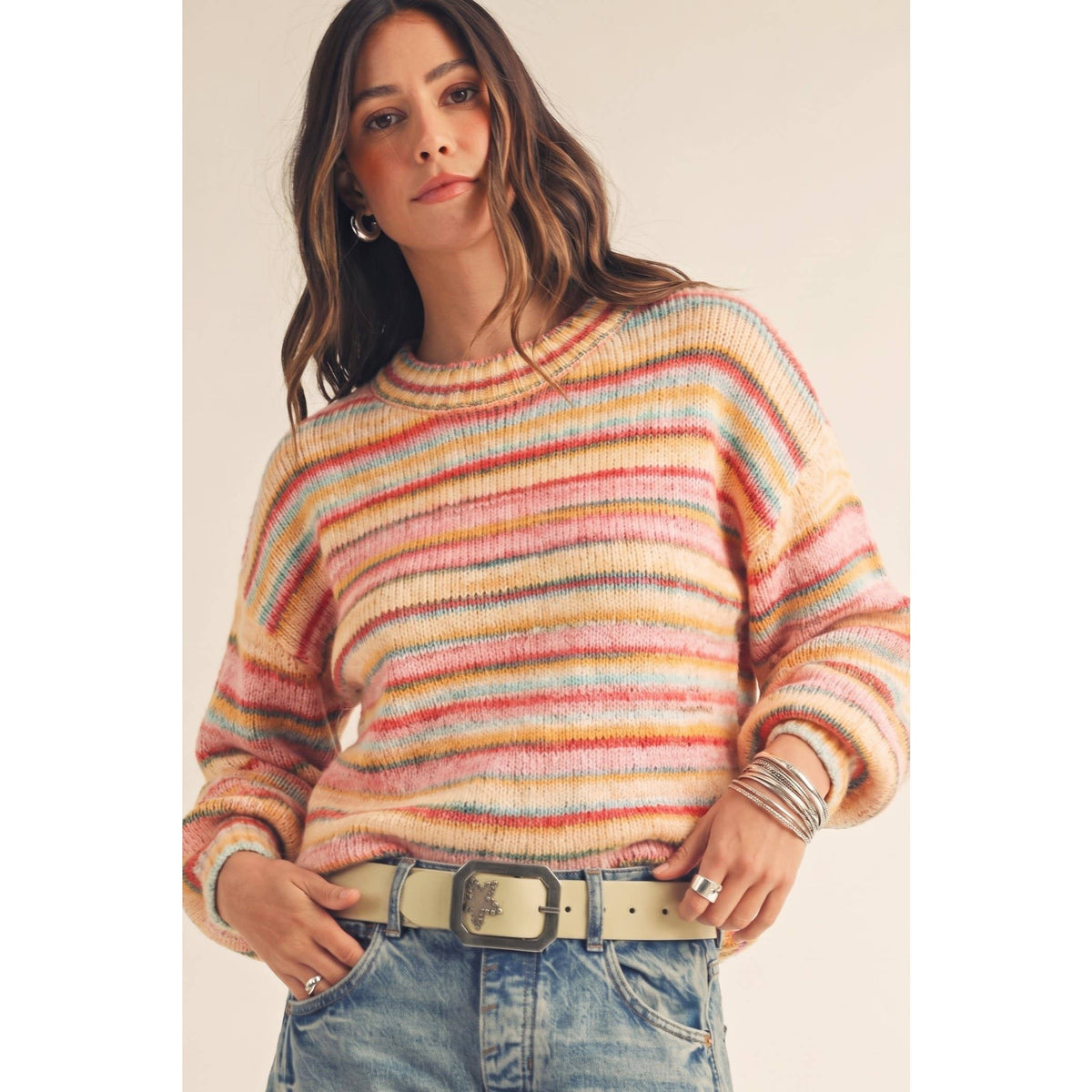 A woman wearing a fuzzy, multicolored striped knit sweater with horizontal bands of pink, orange, yellow, blue, and purple, paired with light-wash blue jeans and a beige belt.