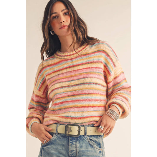 A woman wearing a fuzzy, multicolored striped knit sweater with horizontal bands of pink, orange, yellow, blue, and purple, paired with light-wash blue jeans and a beige belt.