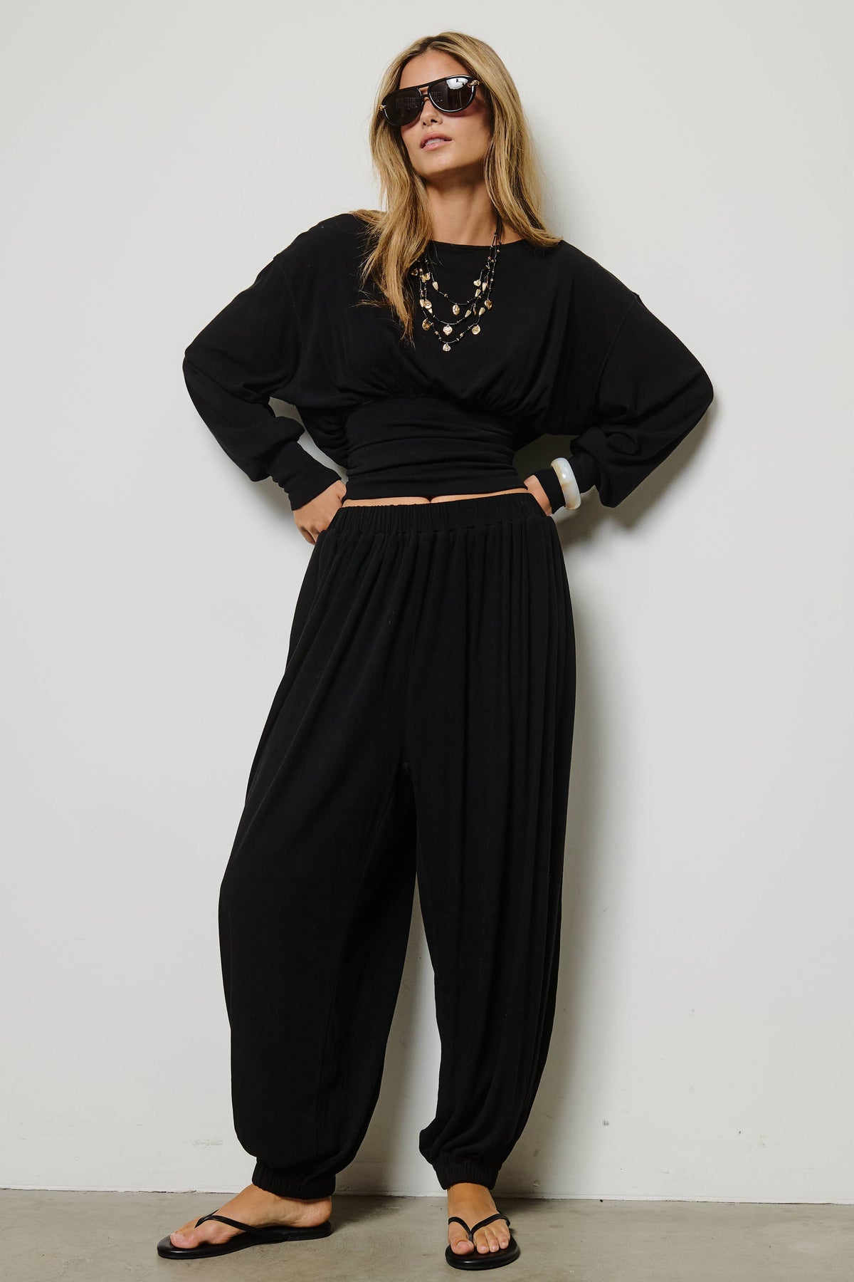 A woman modeling a black two-piece outfit consisting of a long-sleeve wrap-style top and matching loose harem-style pants, styled with sunglasses, layered necklaces, and black flip-flop sandals against a plain light gray wall.
