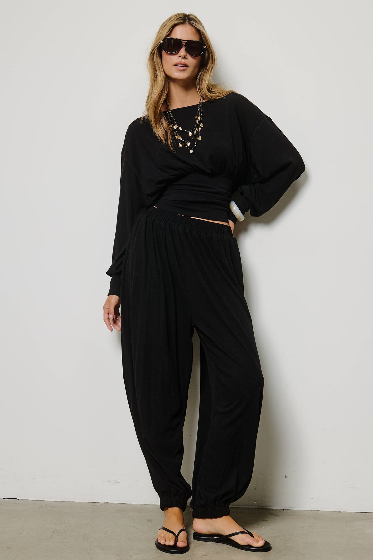 A woman modeling a black two-piece lounge set consisting of a long-sleeve wrap-style top and matching baggy pants, styled with sunglasses, a statement necklace, and black flip-flops against a plain white wall.