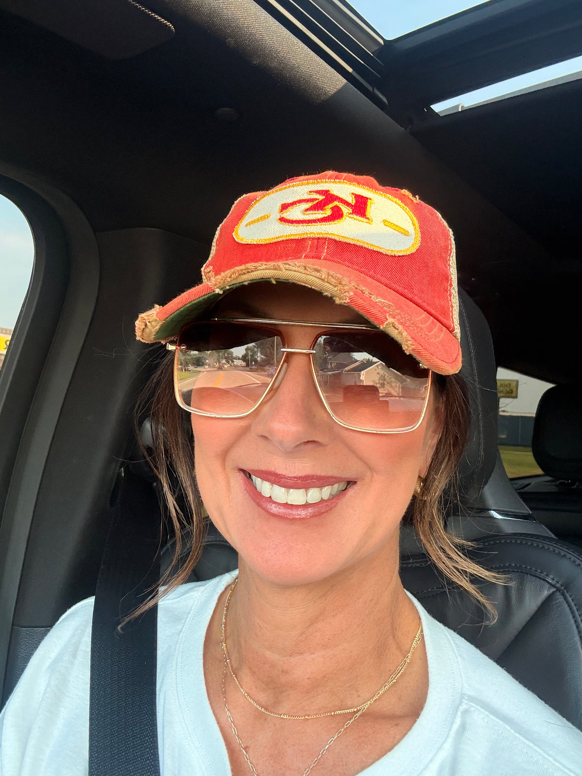 A smiling woman wearing a red distressed baseball cap with a yellow and red logo, gold-framed sunglasses, and a white top, seated inside a car.
