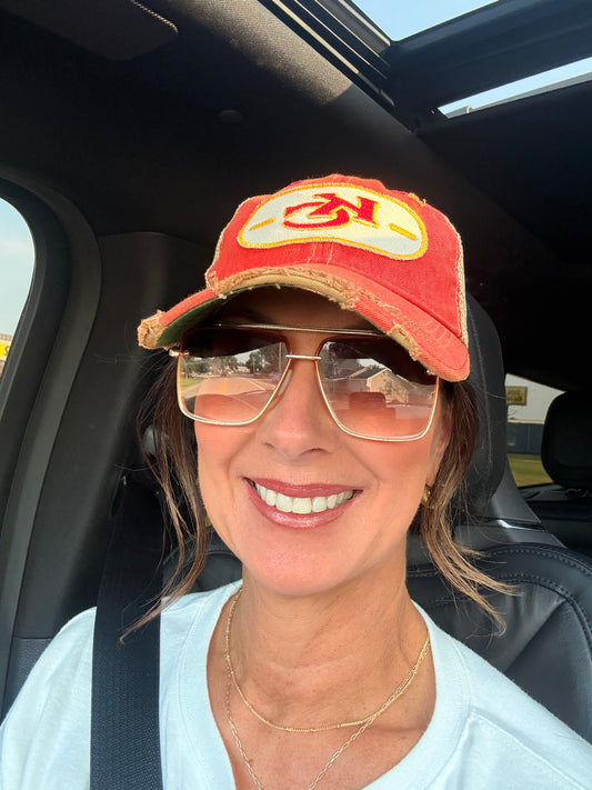 A smiling woman wearing a red distressed baseball cap with a yellow and red logo, gold-framed sunglasses, and a white top, seated inside a car.