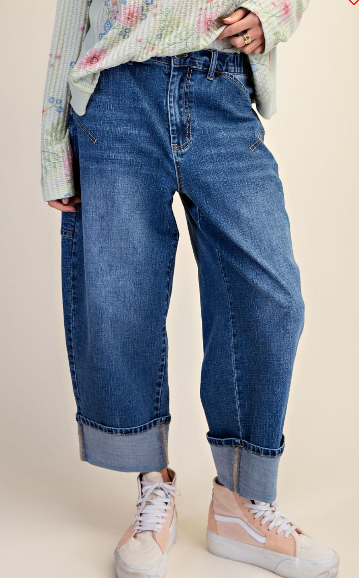 A person wearing loose-fit blue denim jeans with rolled cuffs, styled with a light floral long-sleeve top and pastel sneakers against a plain beige studio background.