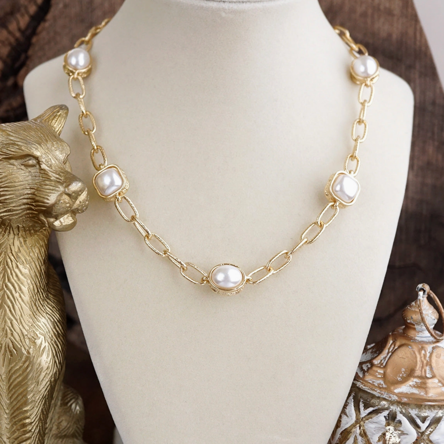 Gold chain necklace with square bezel-set white pearls, displayed on a cream jewelry bust with decorative gold figurines in the background.