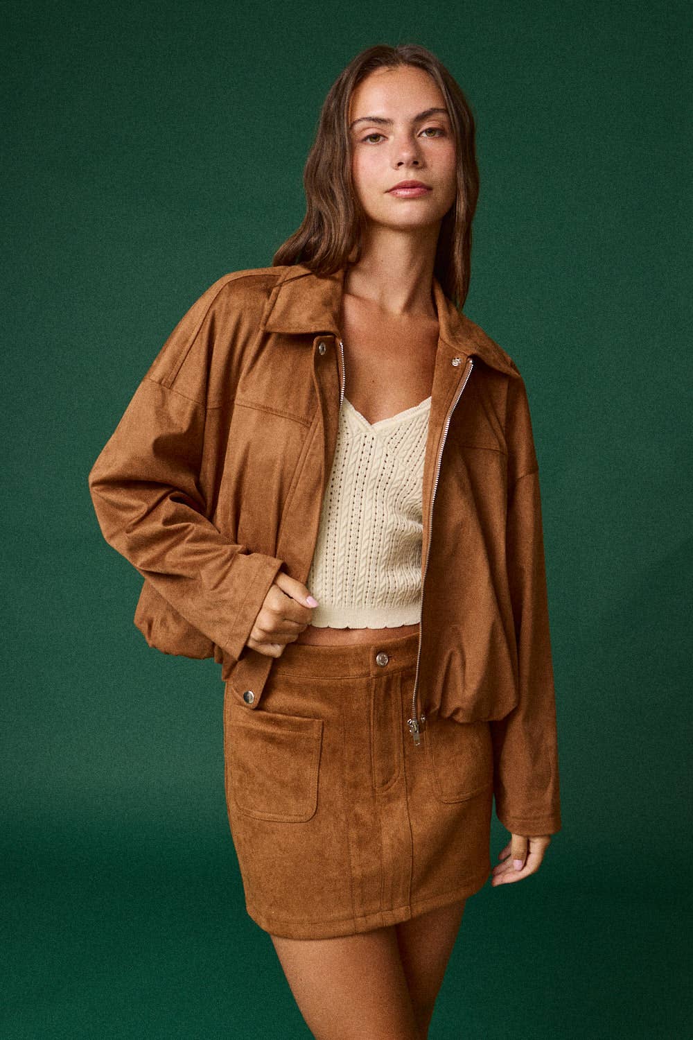 A woman modeling a brown suede jacket and matching mini skirt set, paired with a cream knit top, against a solid dark green studio background.