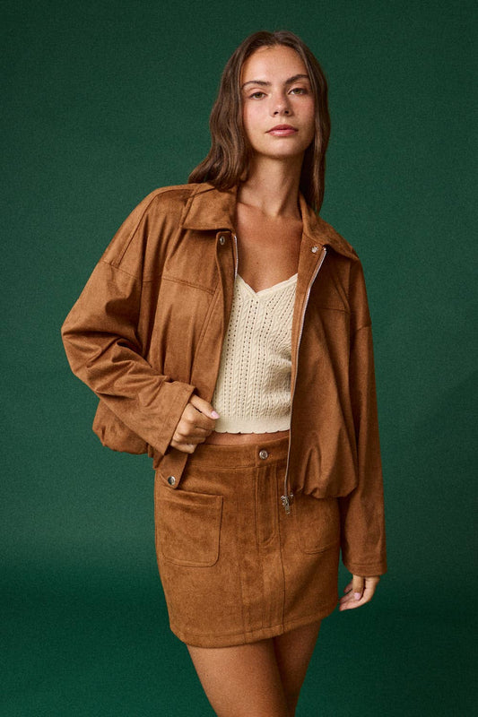 A woman modeling a brown suede jacket and matching mini skirt set, paired with a cream knit top, against a solid dark green studio background.