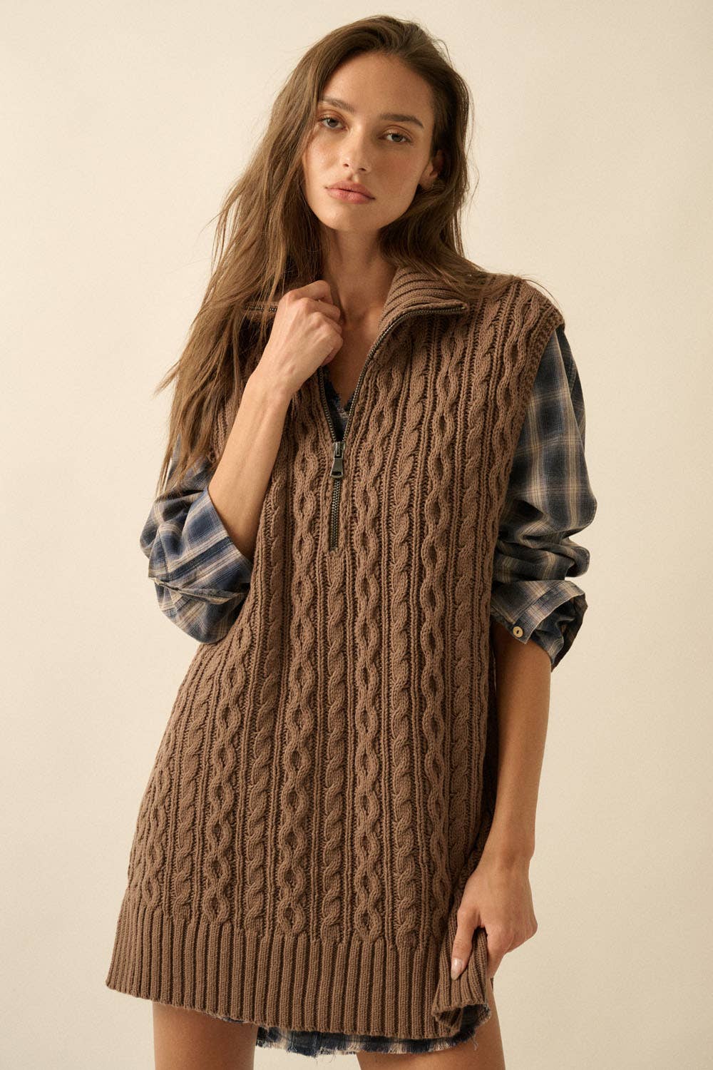 Woman wearing a brown cable knit vest over a plaid shirt on a beige background
