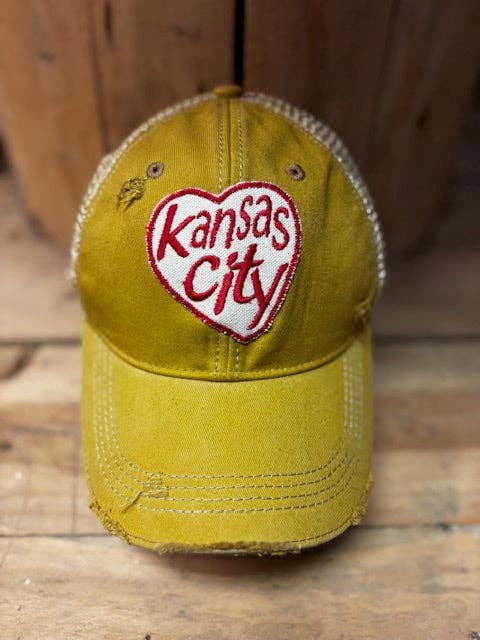 A distressed yellow baseball cap with a white and red embroidered heart patch reading 'Kansas City' on the front, set against a rustic wooden background.